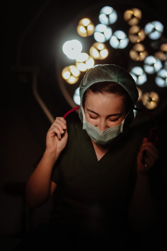 A female surgeon wearing a mask and cap under bright operating room lights.